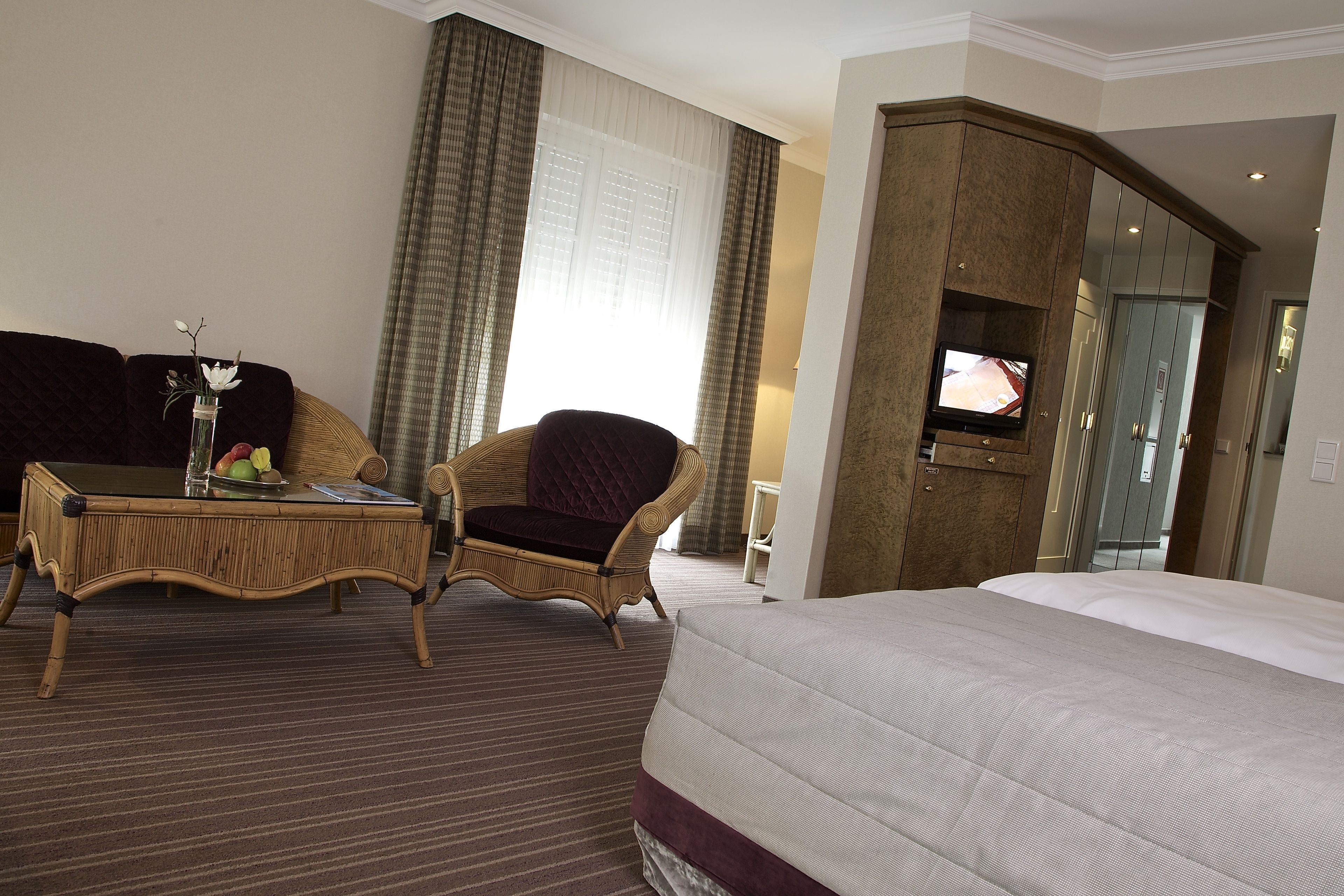 Une chambre d’hôtel avec un grand lit, un fauteuil en osier, une petite table avec un vase et des fruits, et un téléviseur posé sur un meuble.