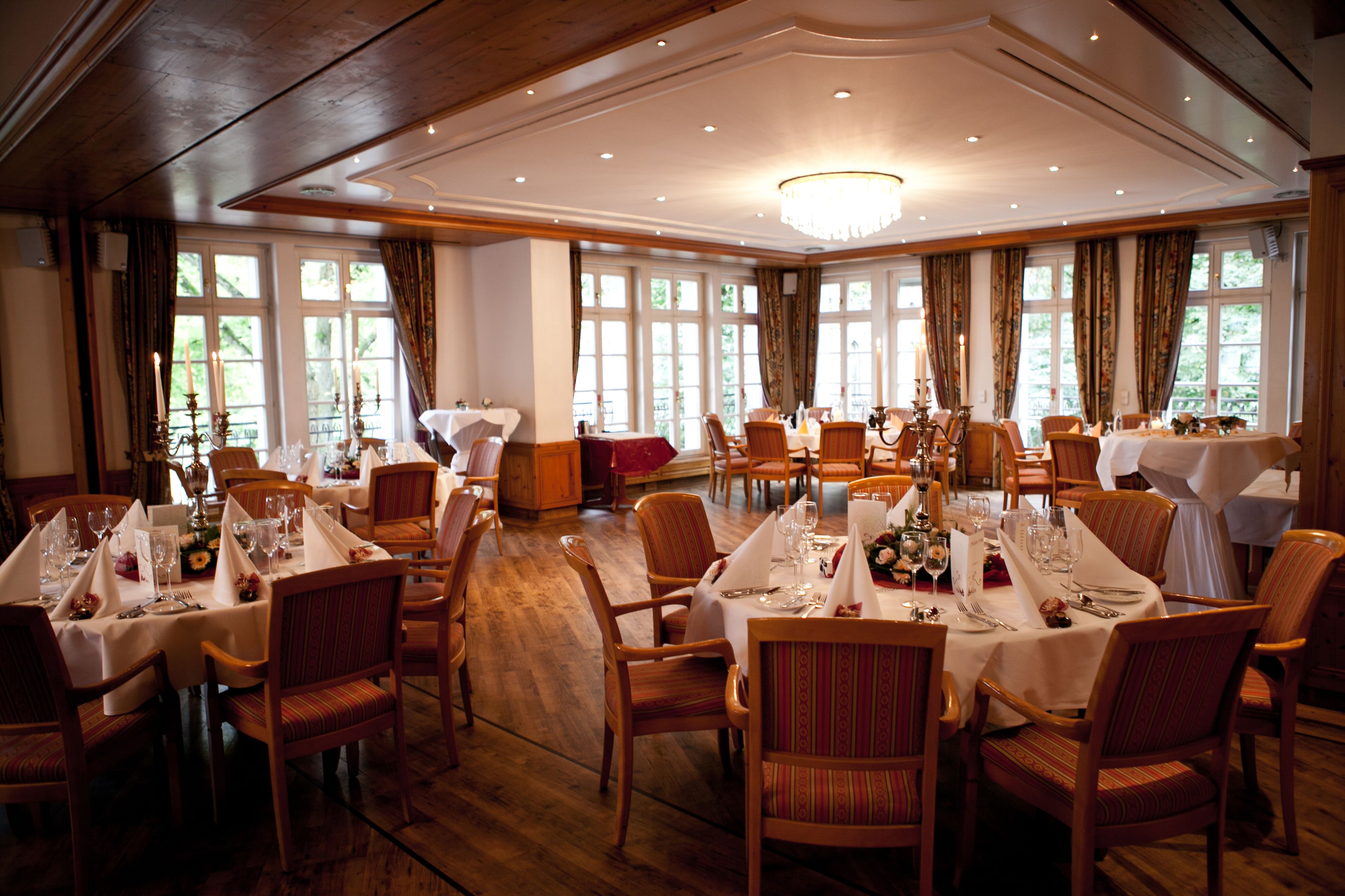 A large dining hall with multiple tables set for a formal meal, wooden floors, and large windows allowing natural light.