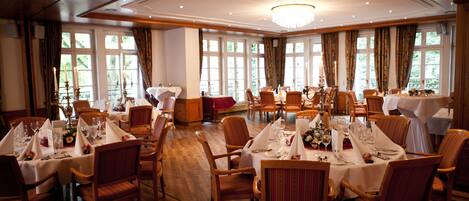 A large dining hall with multiple tables set for a formal meal, wooden floors, and large windows allowing natural light.