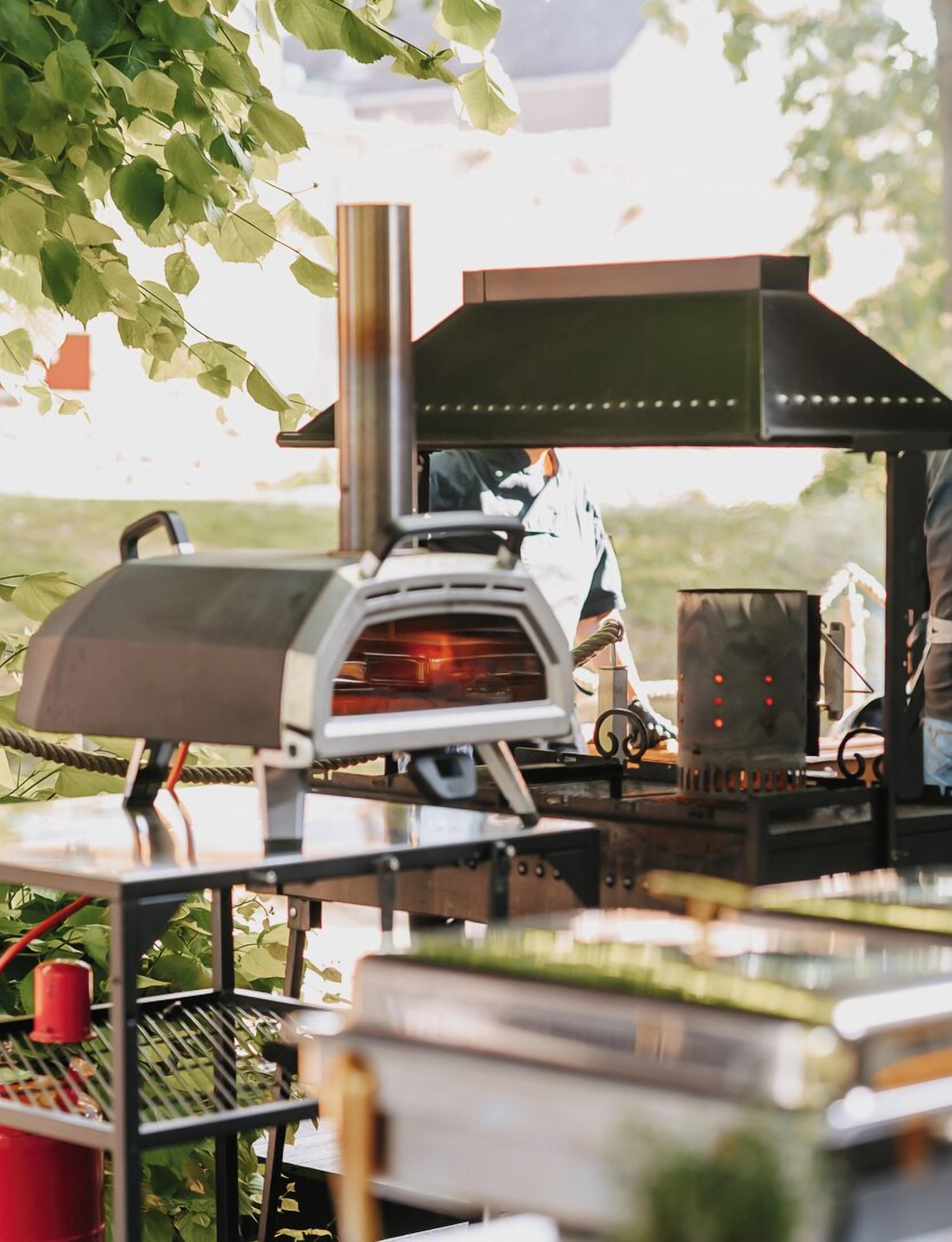 Un espace de cuisson extérieur équipé d’un four à bois, d’un barbecue et d’une personne qui s’en sert.