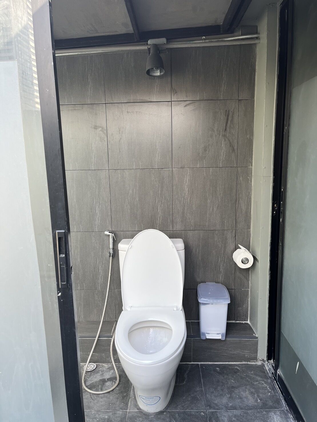 A compact bathroom with a white toilet, a showerhead, and a trash bin.