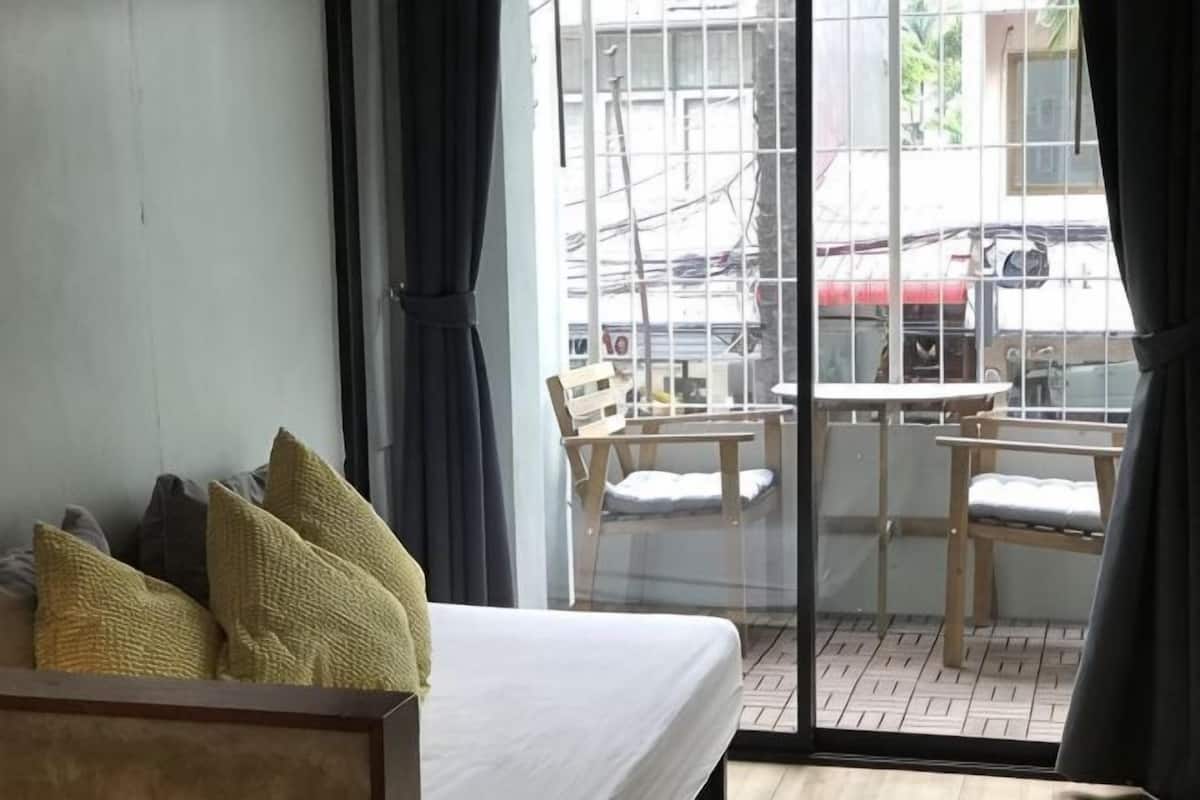A room with a bed, a window with curtains, a balcony with a table and chairs, and a view of the outside.
