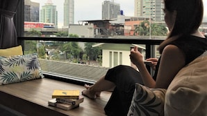 Una persona sentada junto a una ventana con vista a la ciudad, un libro y un almohadón en un repisa de madera.