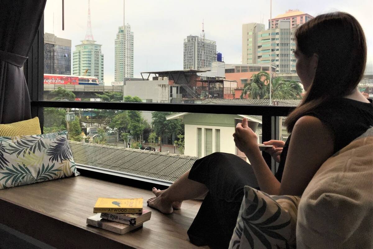 A person sitting by a window with a city view, a book, and a pillow on a wooden ledge.