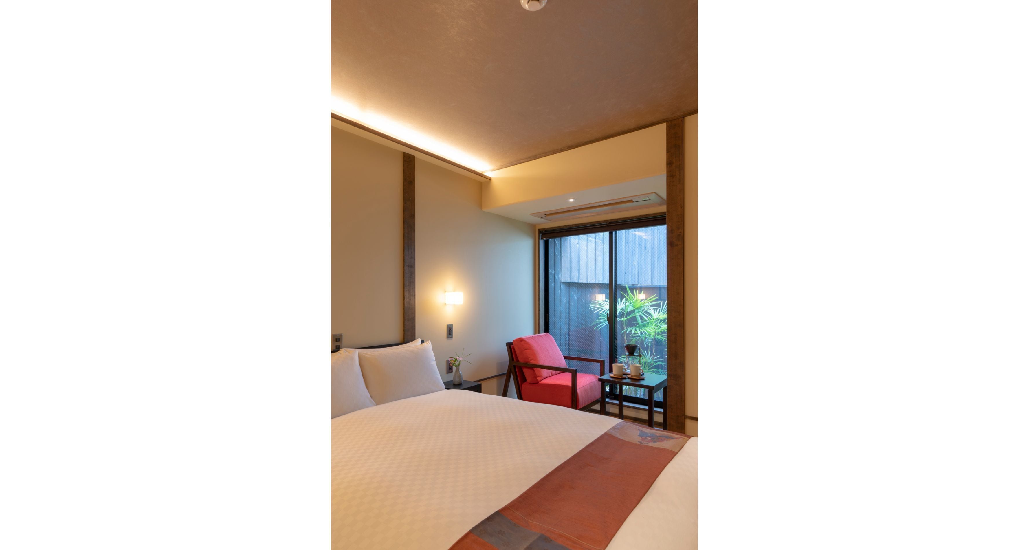 A hotel room with a large bed, a red chair, a small table, and a window with a view of greenery.
