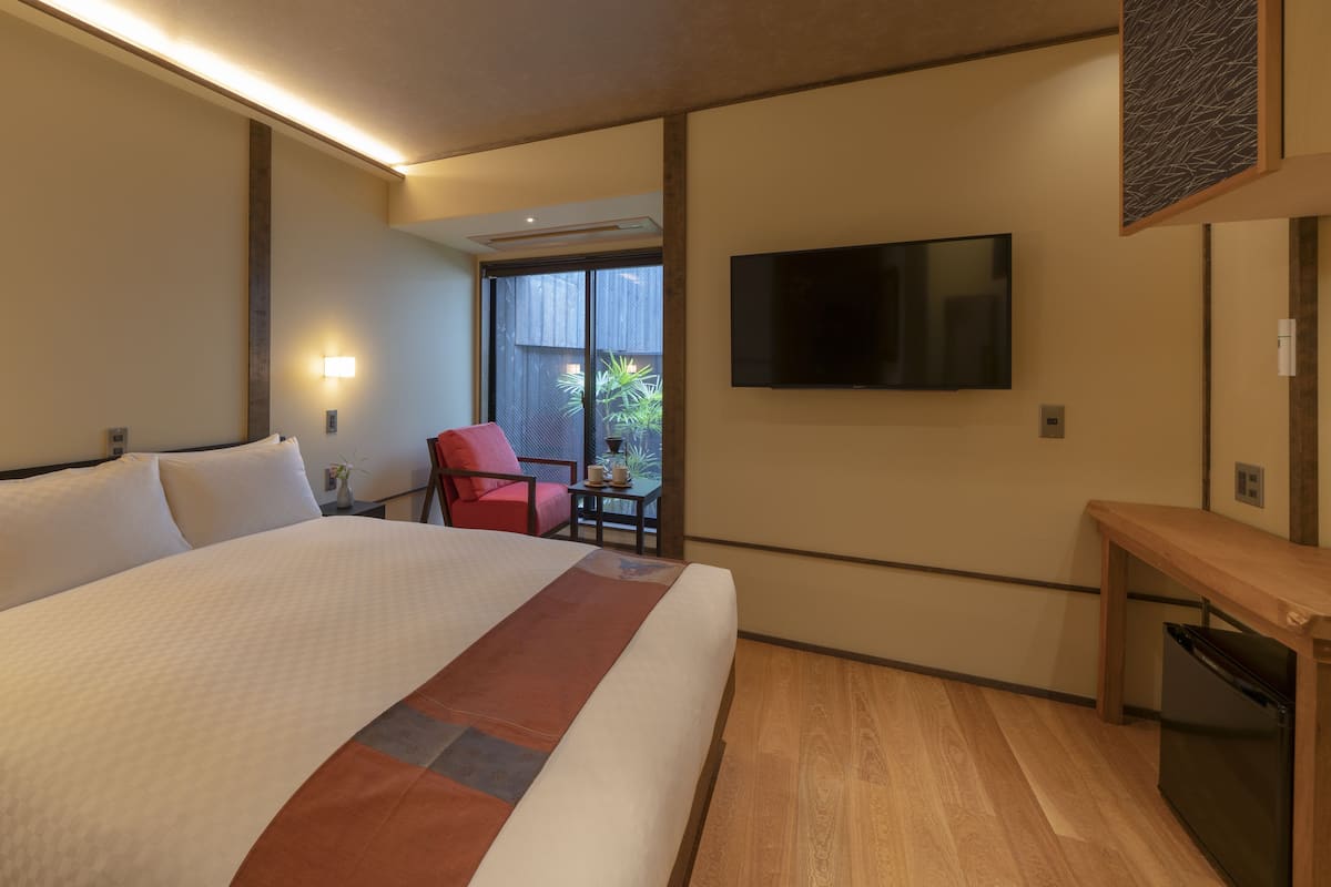 Une chambre d’hôtel avec un grand lit, un téléviseur fixé au mur, une petite table avec une chaise et une fenêtre donnant sur un paysage verdoyant.