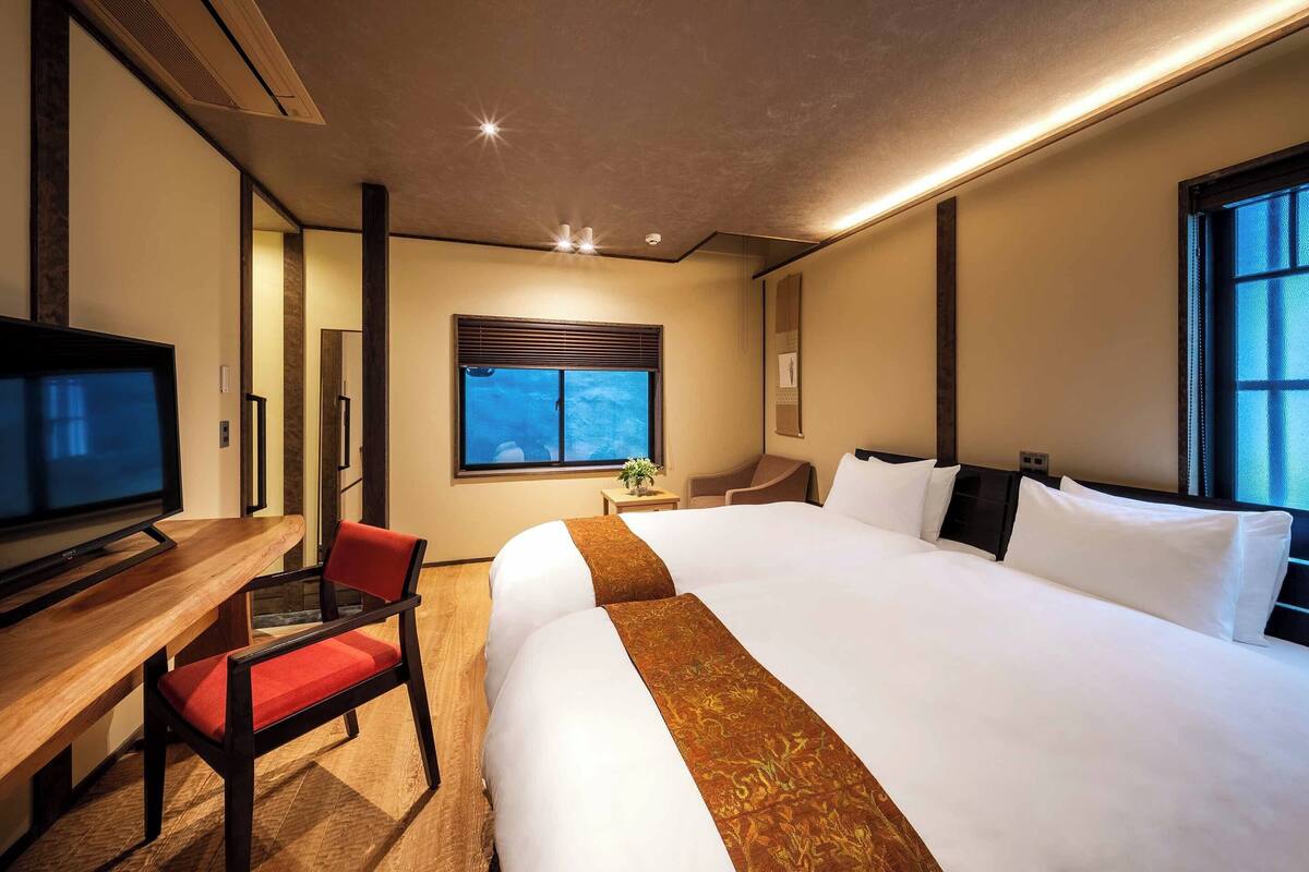 Une chambre d’hôtel avec un grand lit, un bureau avec une chaise, une télévision et une fenêtre avec des stores.