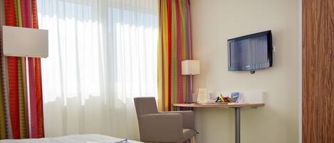 Een hotelkamer met een bed, een bureau met een lamp, een stoel, een televisie aan de muur en kleurrijke gordijnen.