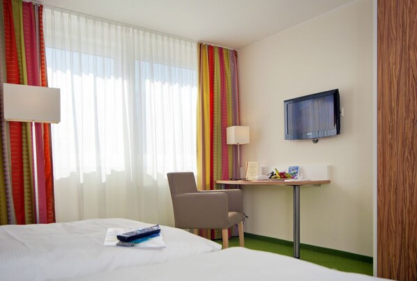 Habitación de hotel con una cama, un escritorio con lámpara, una silla, un televisor en la pared y cortinas coloridas.