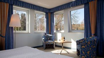 Habitación de hotel con una cama grande, dos sillones, una mesita y una lámpara. Hay tres ventanas con cortinas, y la habitación tiene vistas a árboles.