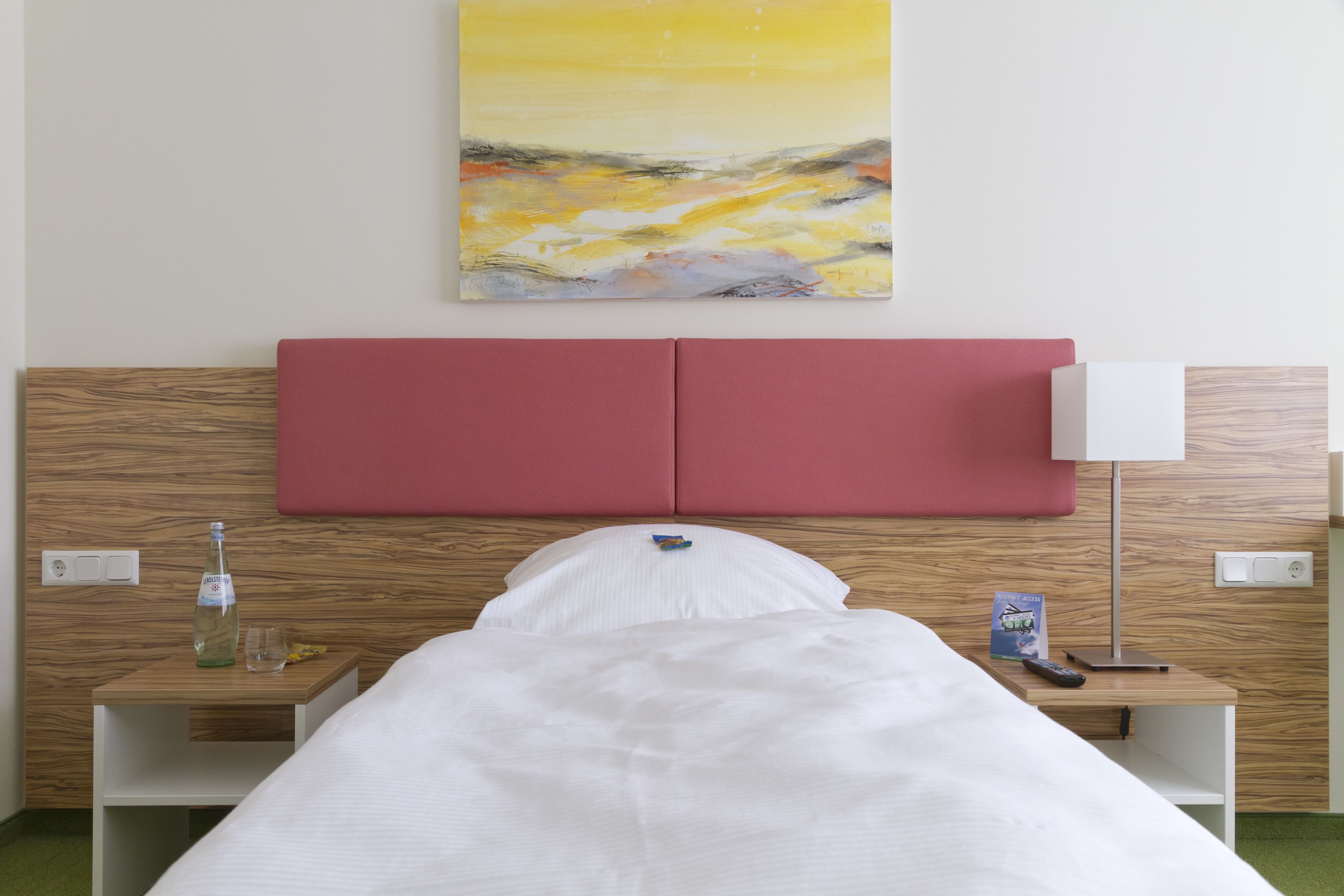 Une chambre d’hôtel avec un lit, une table de chevet sur laquelle se trouvent une bouteille, un verre et un livre, une petite table de nuit avec une lampe, et un tableau accroché au mur.