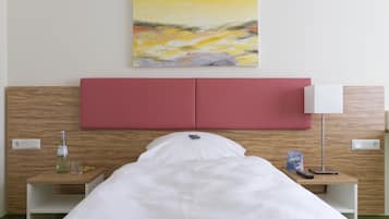 Una habitación de hotel con una cama, una mesita de noche con una botella, un vaso y un libro, una lámpara en la mesita de noche, y un cuadro en la pared.