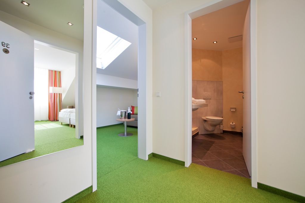 Une chambre d’hôtel avec une moquette verte, un lit, une petite table avec une bouteille, et une fenêtre donnant sur l’extérieur.