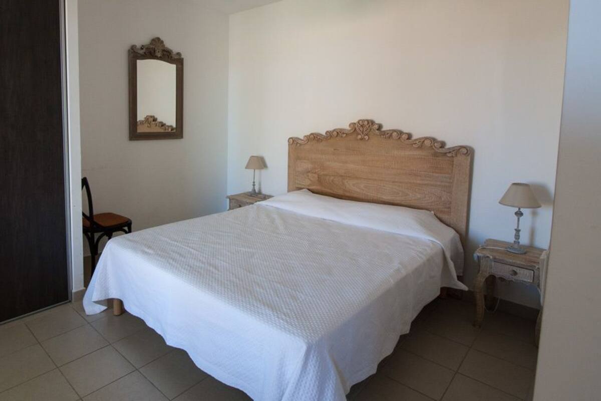 Un dormitorio con una cama grande, cabecera de madera, dos mesitas de noche con lámparas, una silla y un espejo en la pared.