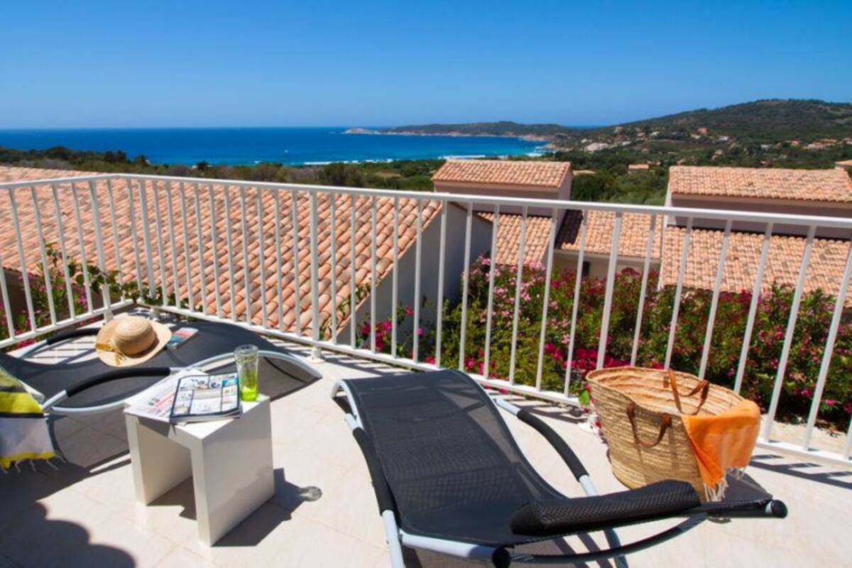 Un balcón con vista al mar, una silla, una mesa con un sombrero y una cesta.