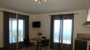 A modern living room with a sofa, a dining table with chairs, a TV mounted on the wall, and a kitchen area with a stove. - Résidence Castugna (Cargese)