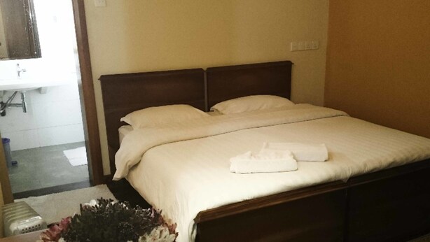 A neatly made bed with white linens and a folded towel on the bed. There is a wooden headboard, a small table with a vase of flowers, and a patterned rug on the floor.