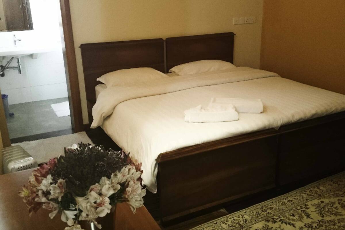 A neatly made bed with white linens and a folded towel on the bed. There is a wooden headboard, a small table with a vase of flowers, and a patterned rug on the floor.