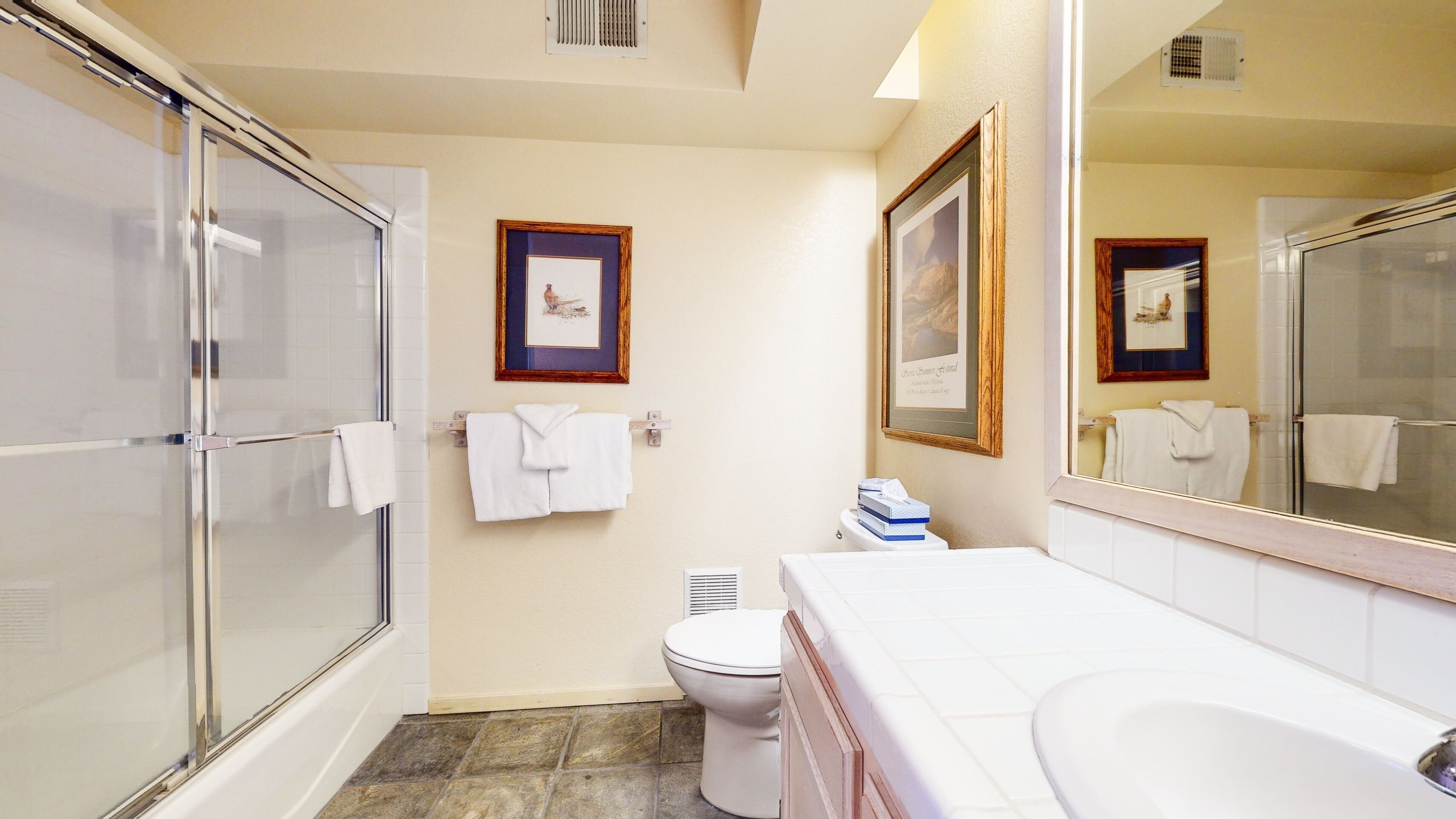 A bathroom with a glass shower enclosure, a toilet, a sink with a mirror, and two framed pictures on the wall.