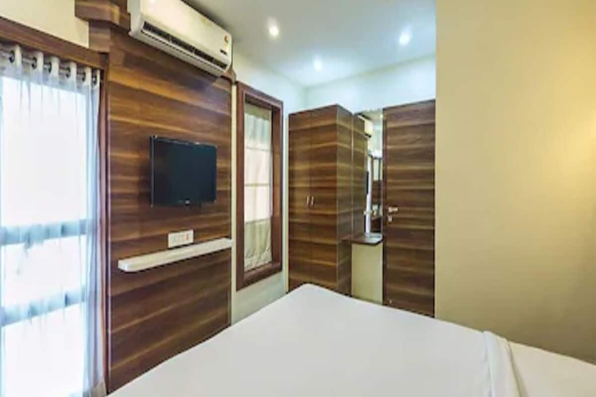 A hotel room with a bed, a TV mounted on the wall, a window with curtains, and wooden paneling on the walls.