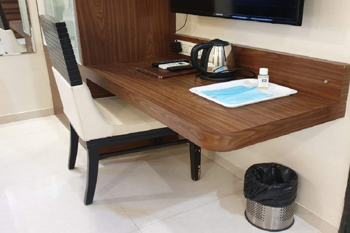 A hotel room with a wooden desk, a television mounted on the wall, a trash bin, and a chair.