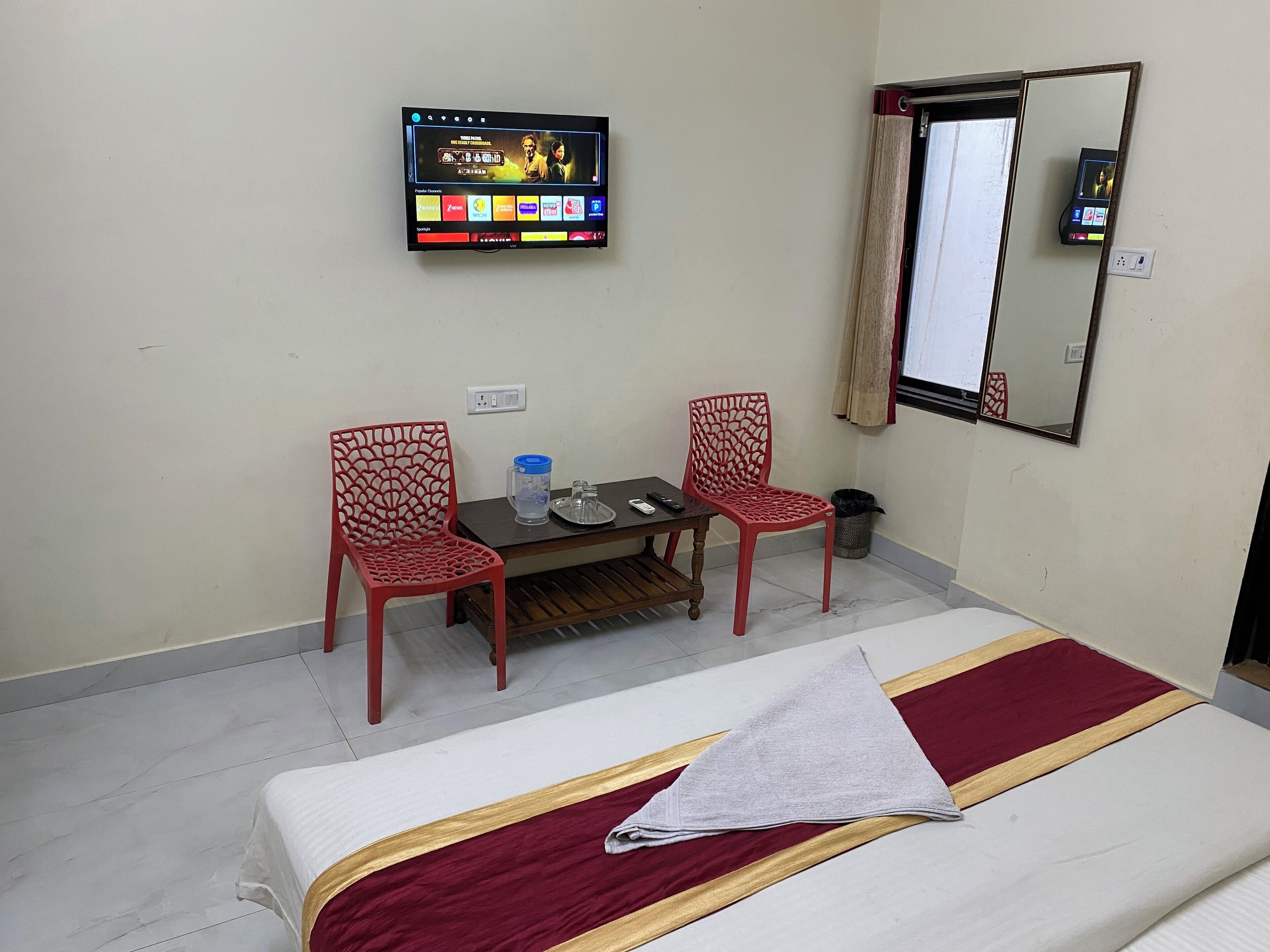 A hotel room with a bed, two red chairs, a small table with a water jug, a TV mounted on the wall, and a mirror.