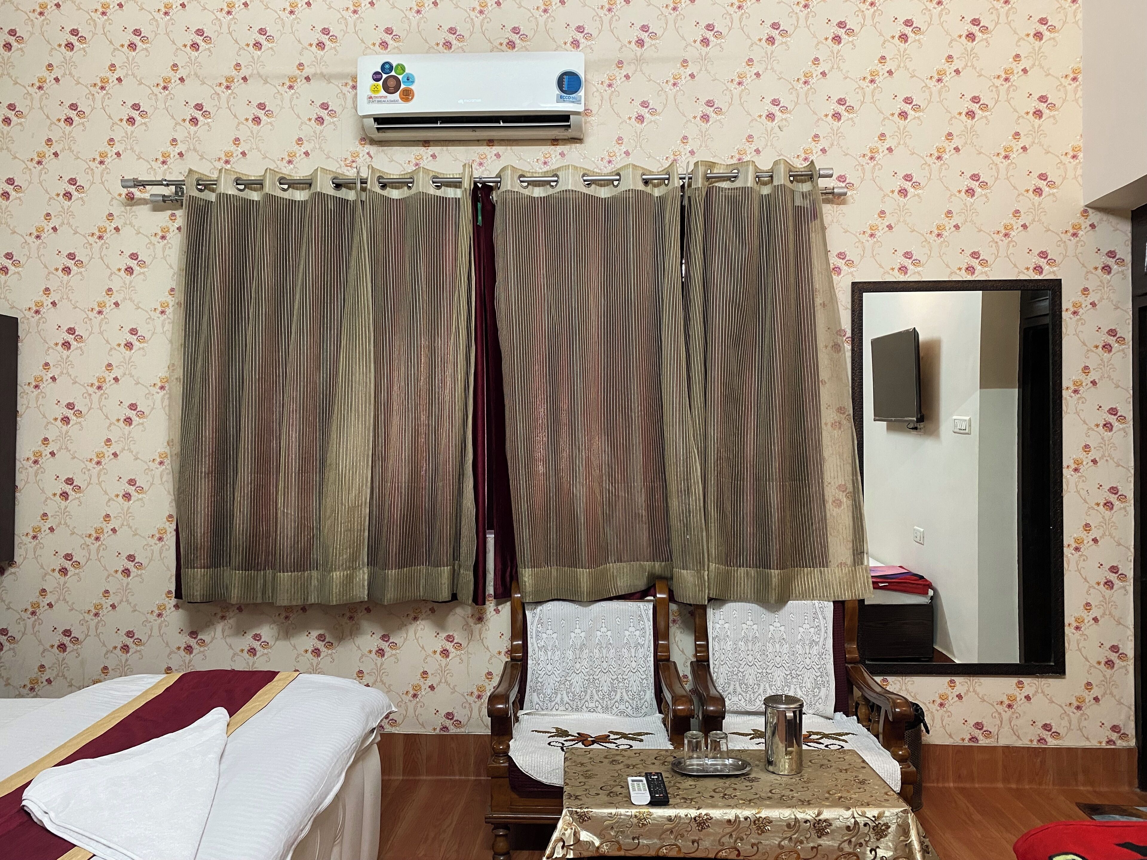 Une chambre d’hôtel comprenant un lit, une table avec un plateau, une chaise, une chaise en bois, un climatiseur fixé au mur et un miroir.