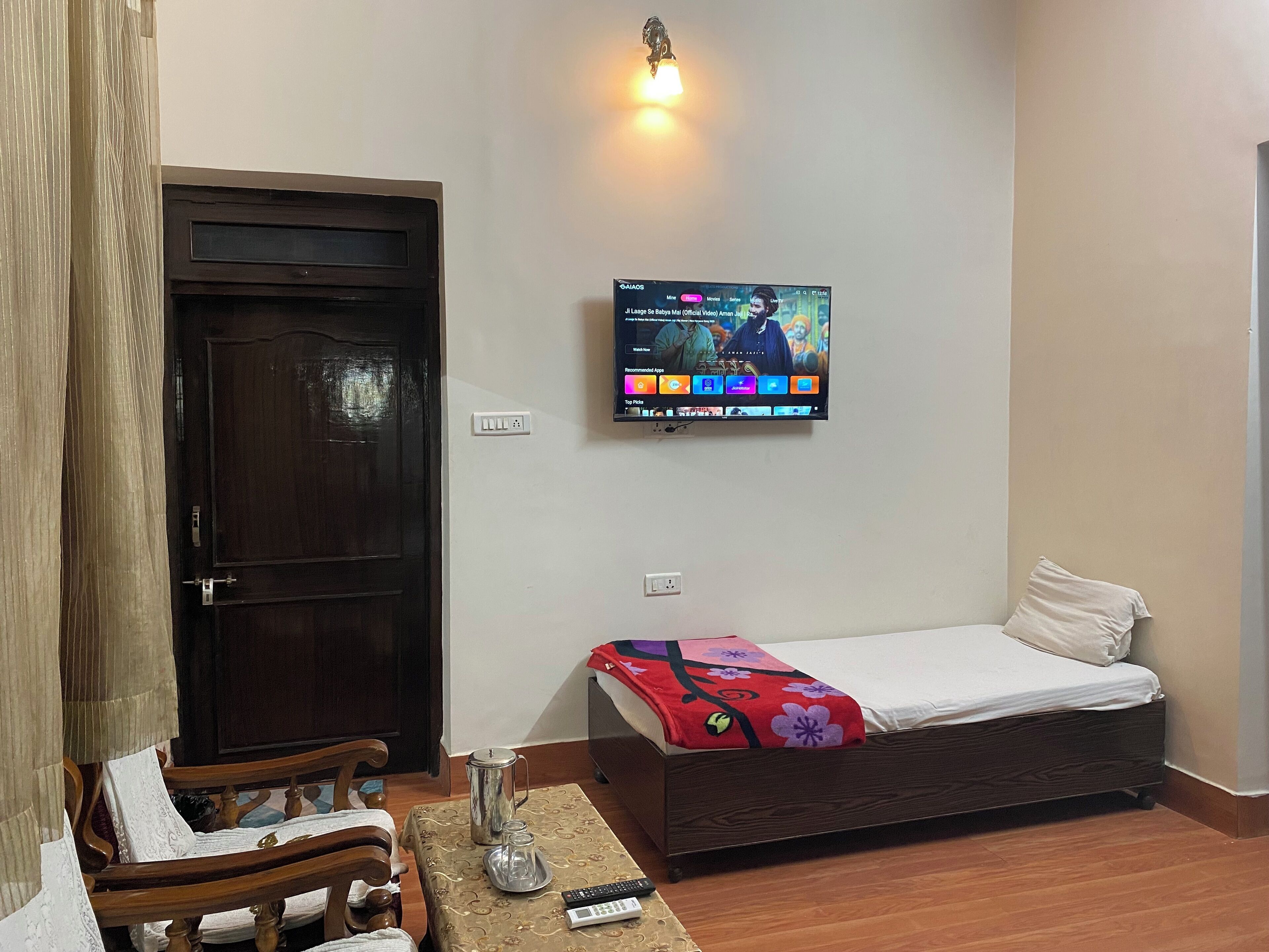 A room with a bed, a television mounted on the wall, a wooden door, and a small table with a remote control.