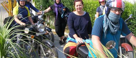 A group of people on motorcycles, some wearing helmets, in an outdoor setting with greenery and buildings in the background.