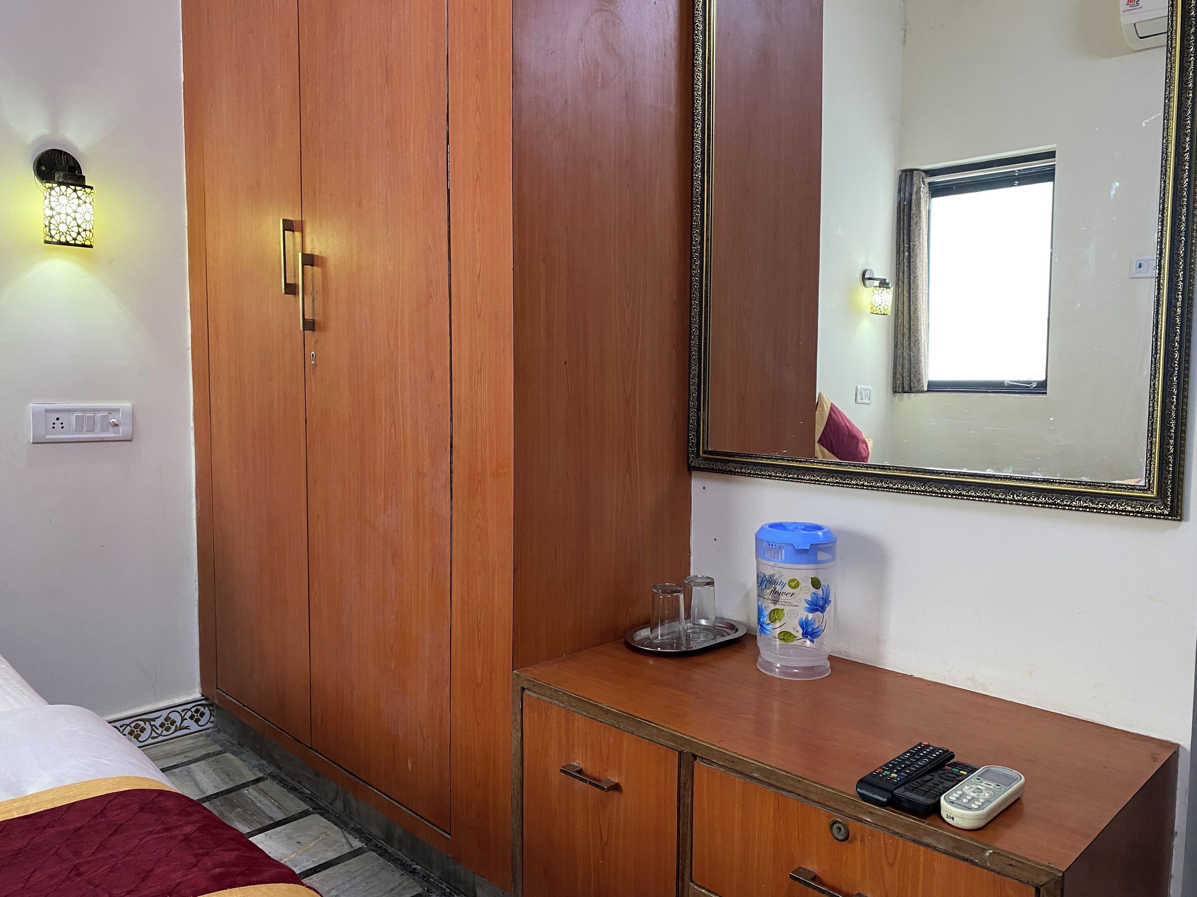 A hotel room with a wooden wardrobe, a mirror, a bedside table with a water bottle, and a bed with a red and gold bedspread.