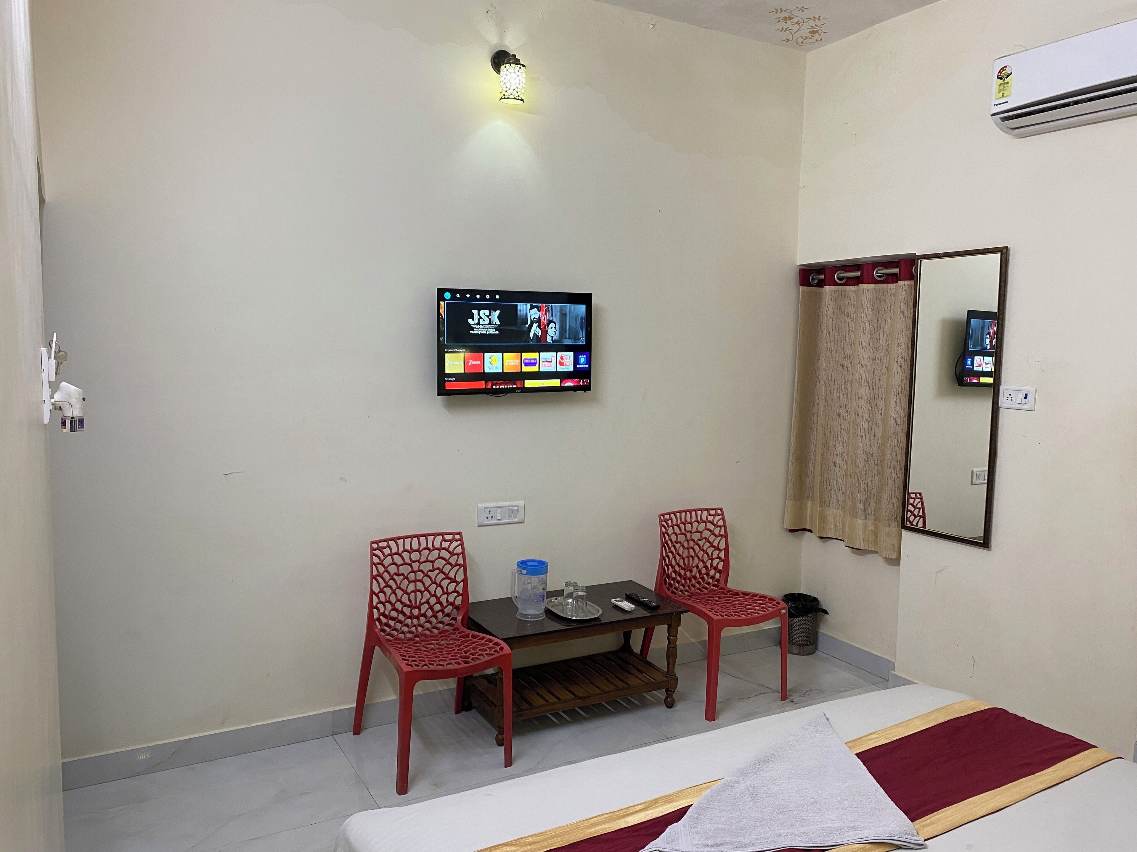 A hotel room with a bed, two red chairs, a small table with a bottle and remote control, a TV mounted on the wall, and a mirror.