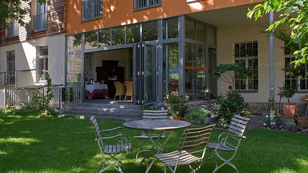 A patio with a table and chairs, a glass door entrance, and a garden with plants and a wooden bench. - Alte Remise (Dresden)