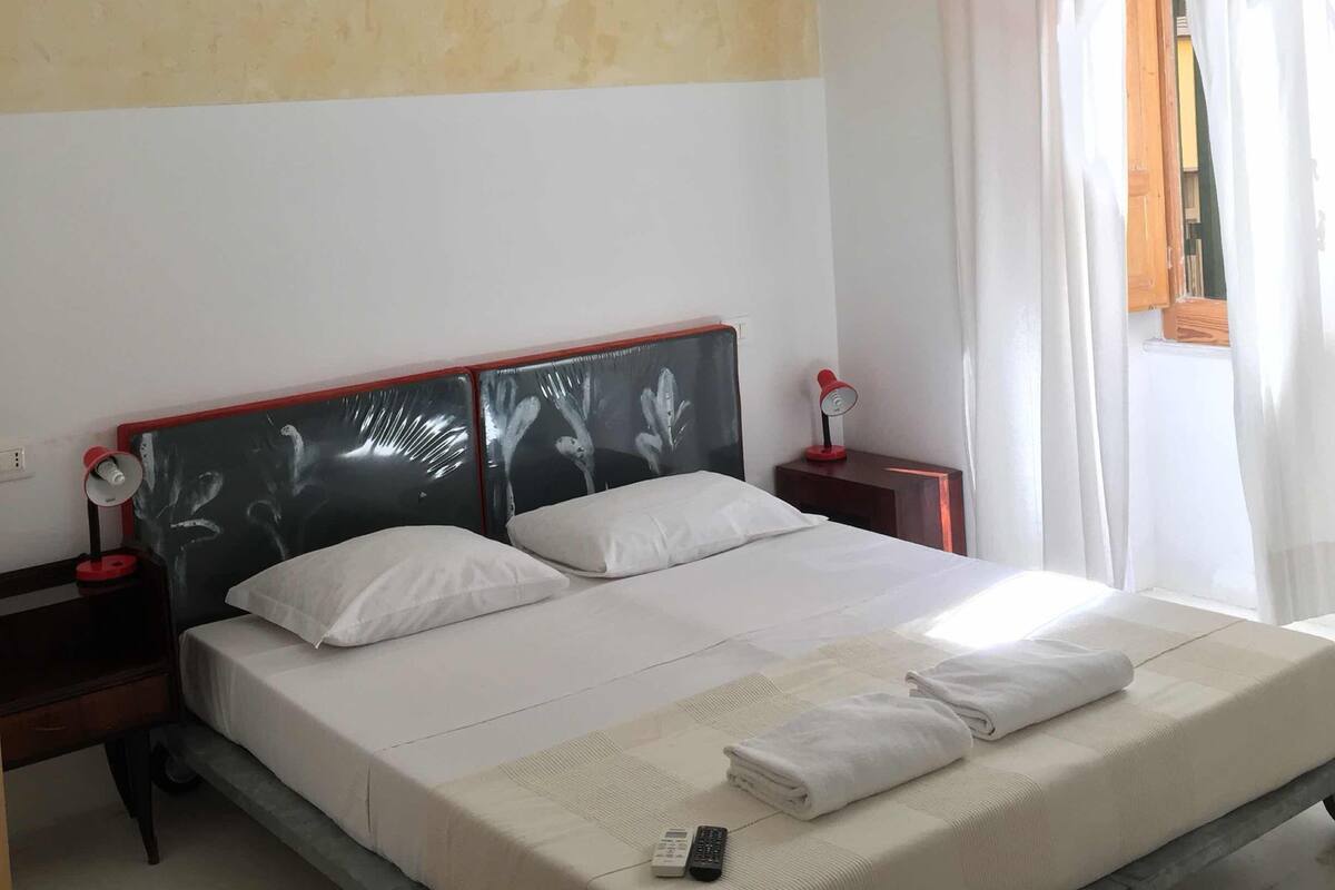 A hotel room with a bed, a bedside table, a nightstand, a wall-mounted light, and a window with curtains.