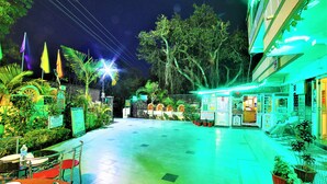 A spacious outdoor area with a tiled floor, surrounded by greenery and potted plants. There are tables and chairs set up for dining, and a building with signage in the background. - Shri Ganesh Hotel (Abu Road)