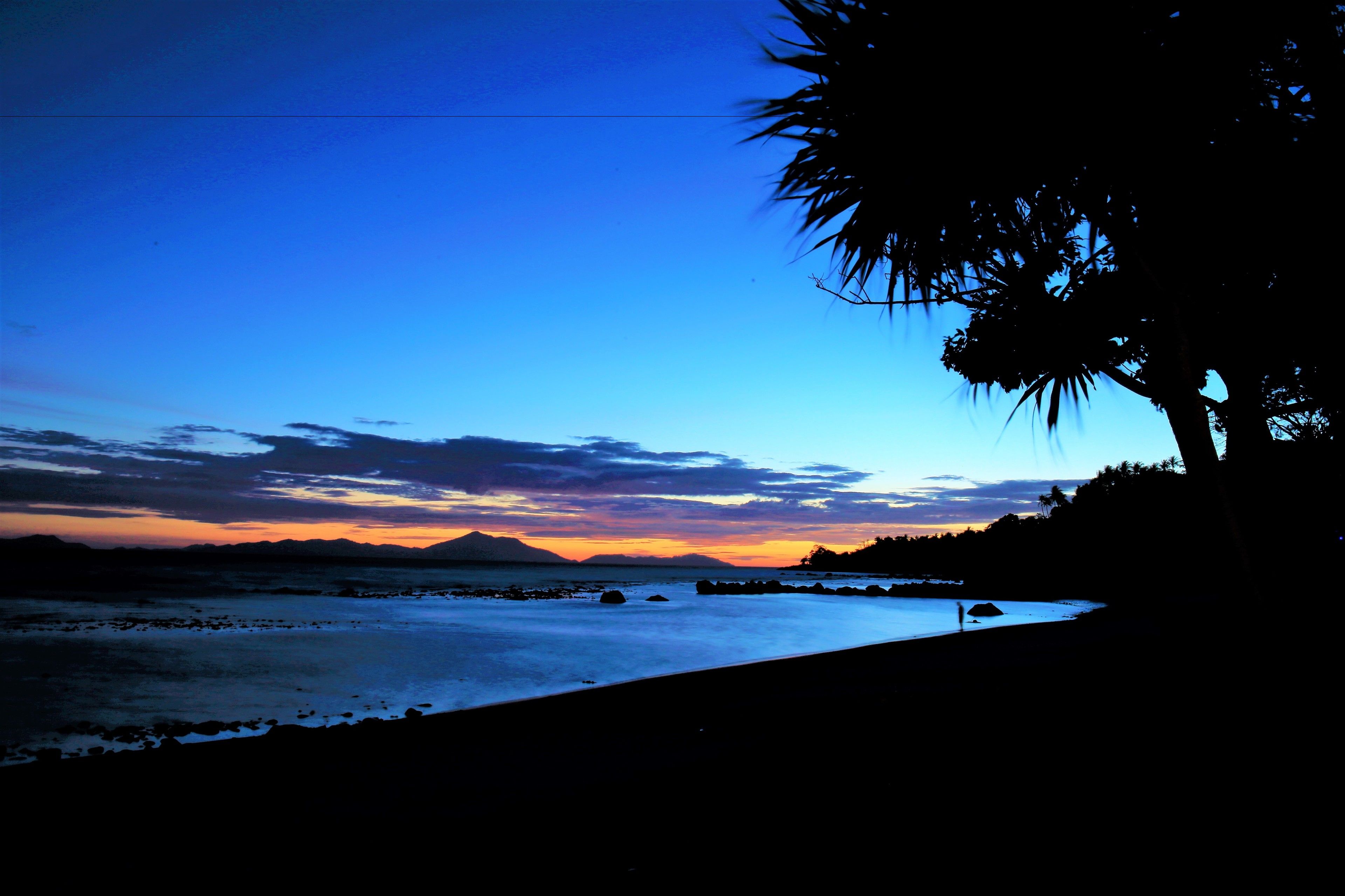 A beach at sunset with silhouettes of trees and distant mountains.