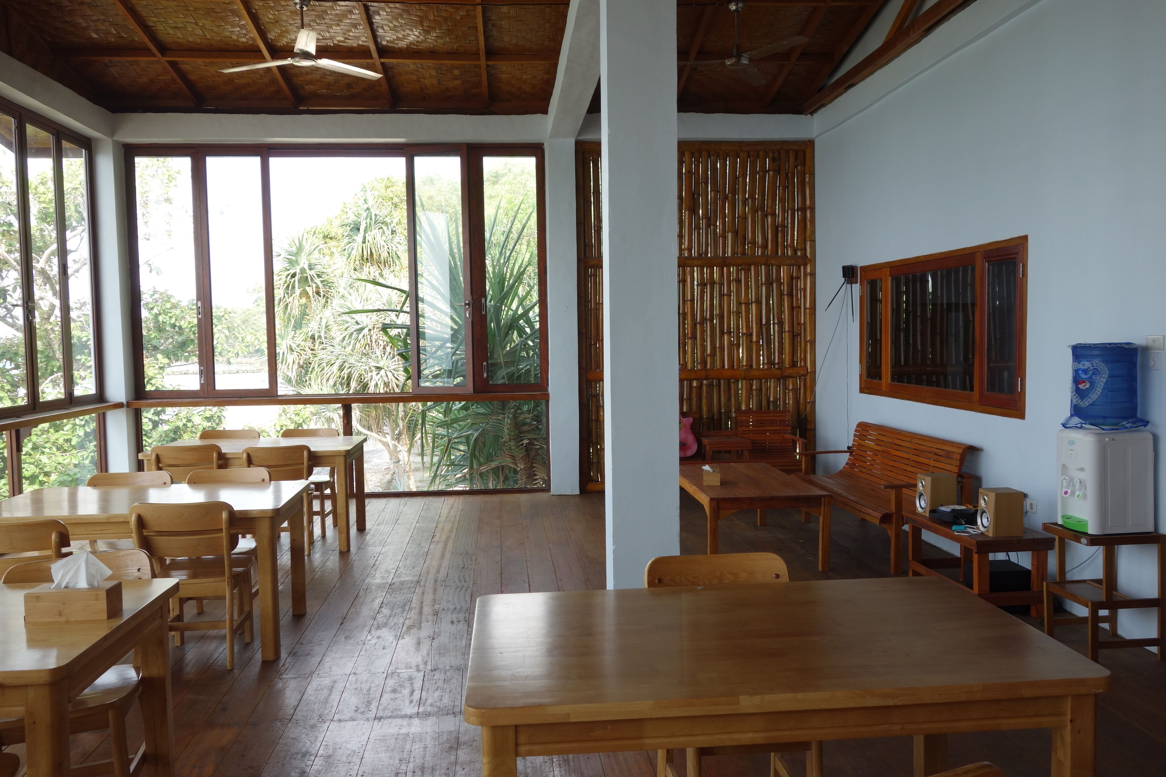A spacious room with wooden tables and chairs, large windows with a view of greenery, a ceiling fan, and a water dispenser.