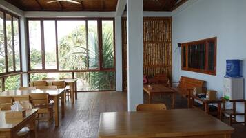 A spacious room with wooden tables and chairs, large windows with a view of greenery, a ceiling fan, and a water dispenser.