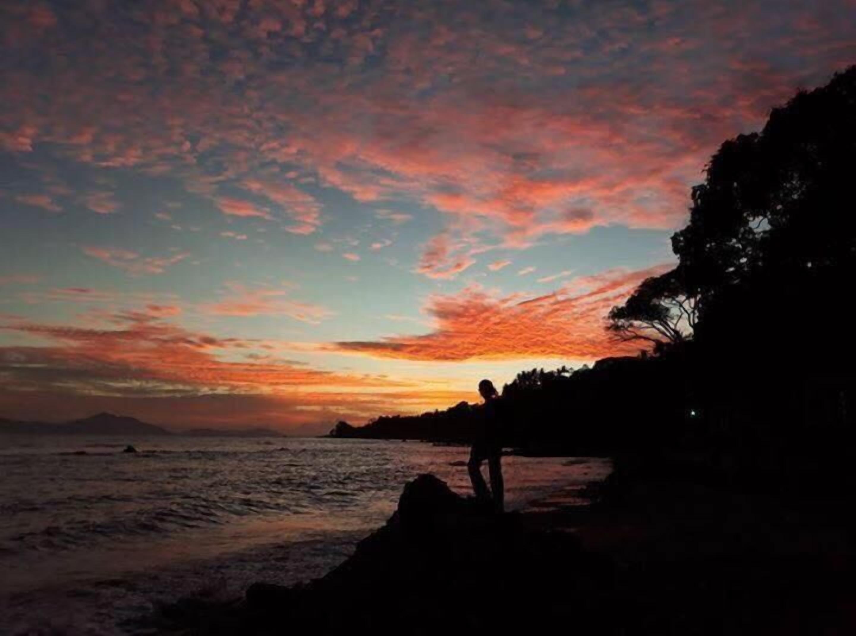 A sunset over a coastal landscape with a silhouette of a person standing on rocks.