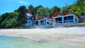 A beachfront property with multiple wooden-roofed buildings, a sandy beach, and lush greenery in the background.
