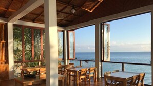 A spacious dining area with wooden tables and chairs, large windows offering a view of the sea, and a wooden ceiling.