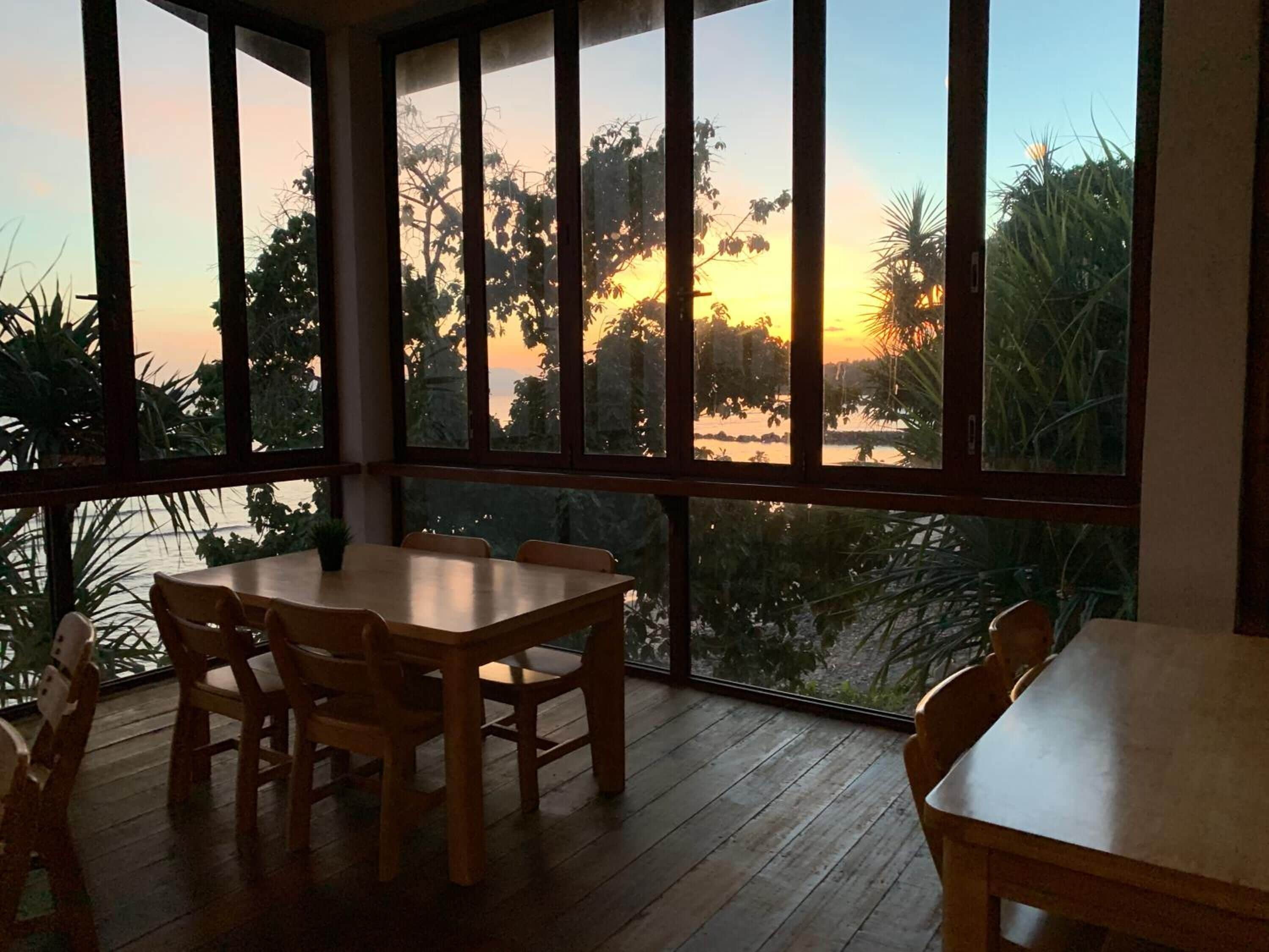 A room with a large window overlooking a body of water and trees, wooden furniture, and a view of the sunset.