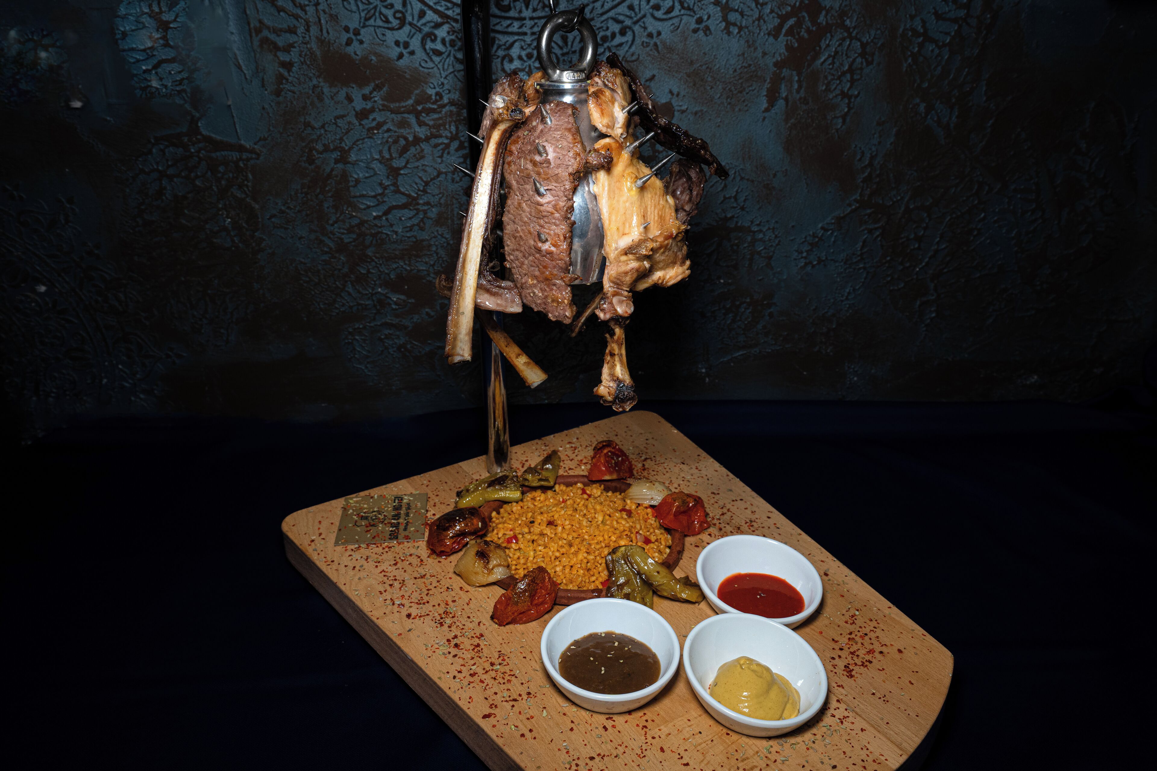A roasted meat dish with vegetables and two dipping sauces on a wooden board.