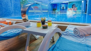 Zona de piscina con sillones reclinables, una mesa con bebidas y aperitivos, y una piscina cubierta con vidrio.