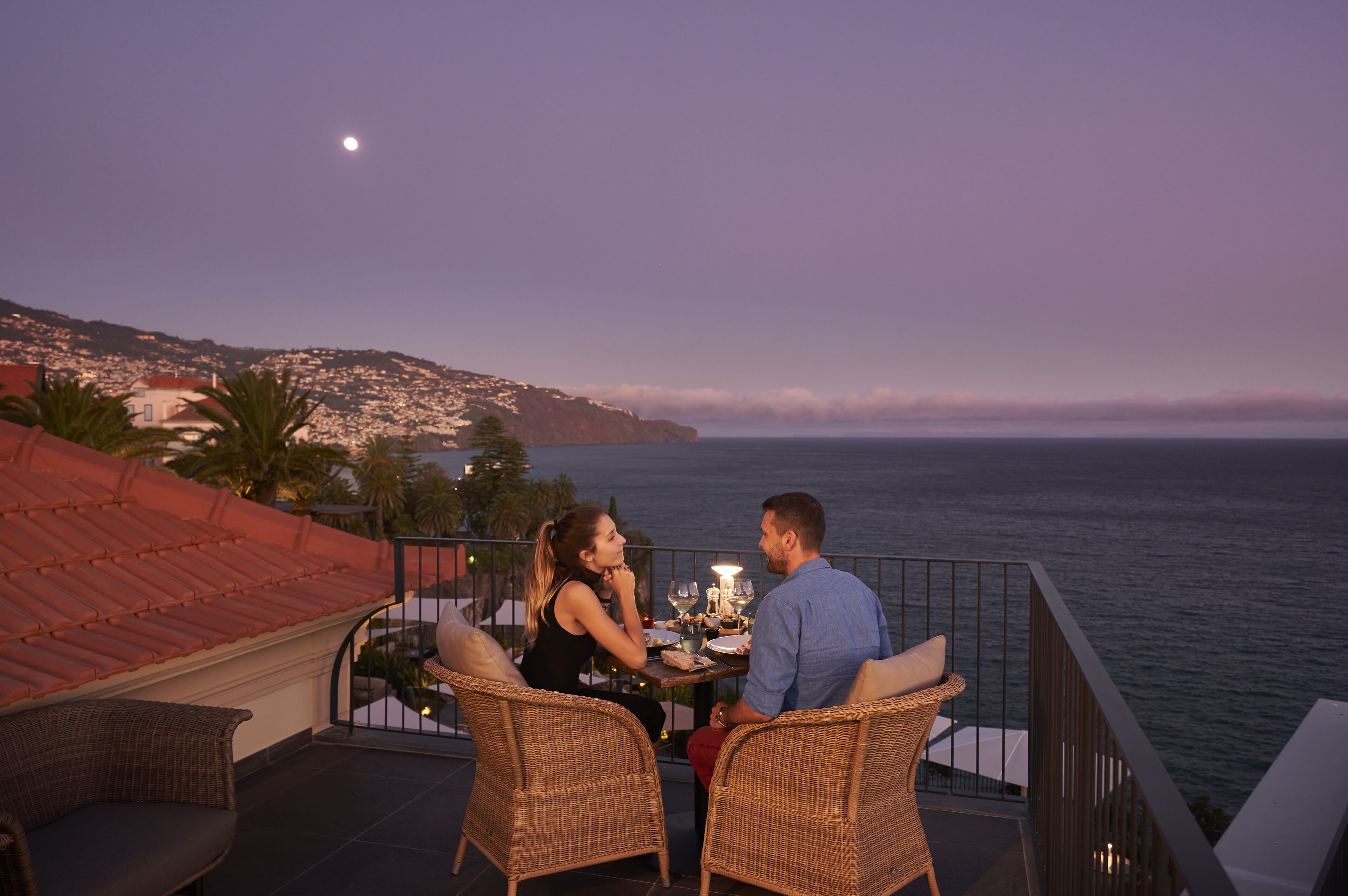 A couple dining at a table with a view of the sea and a mountain in the background.