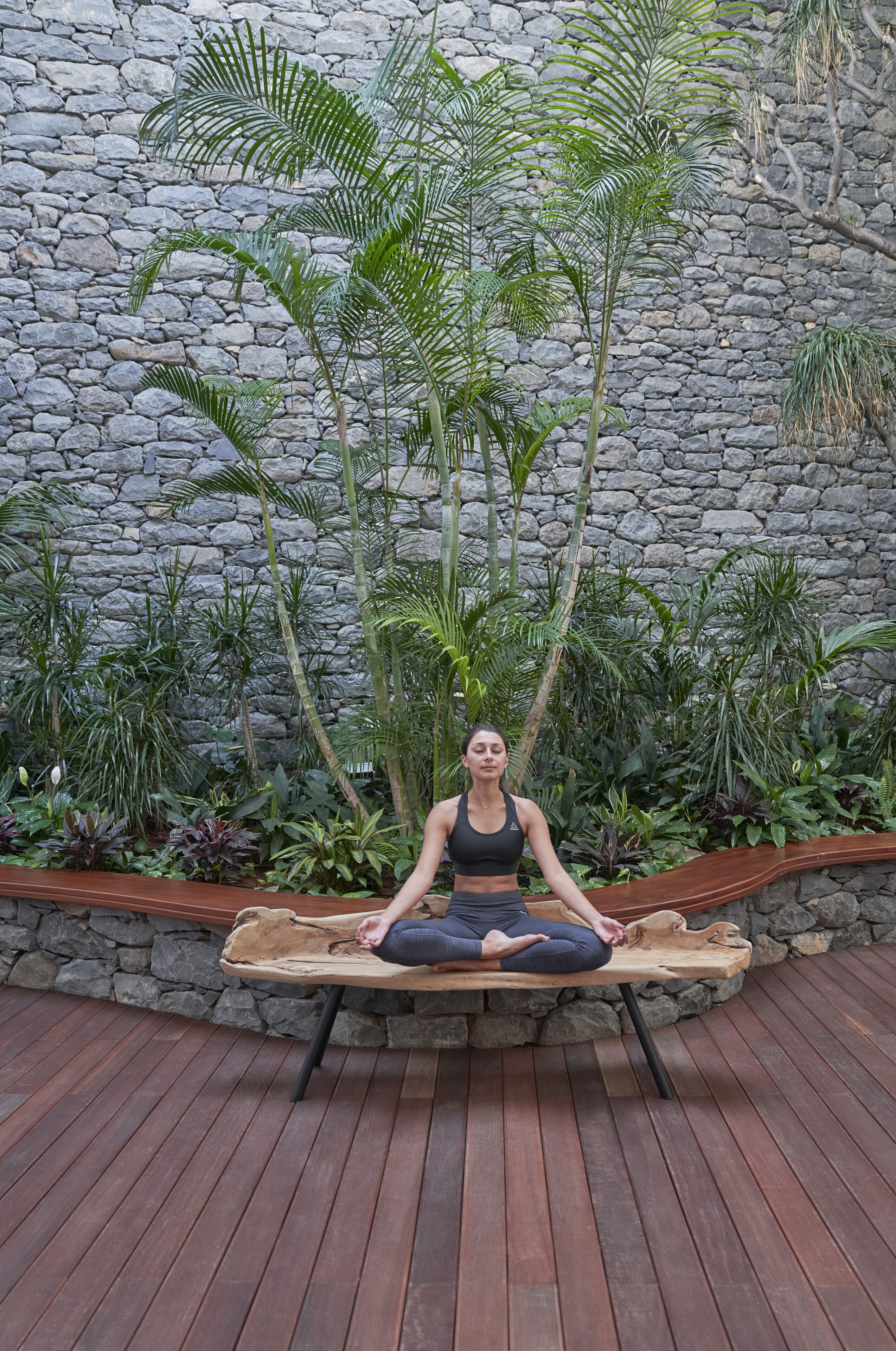 A person meditating on a wooden bench in an outdoor setting with a stone wall and tropical plants in the background.