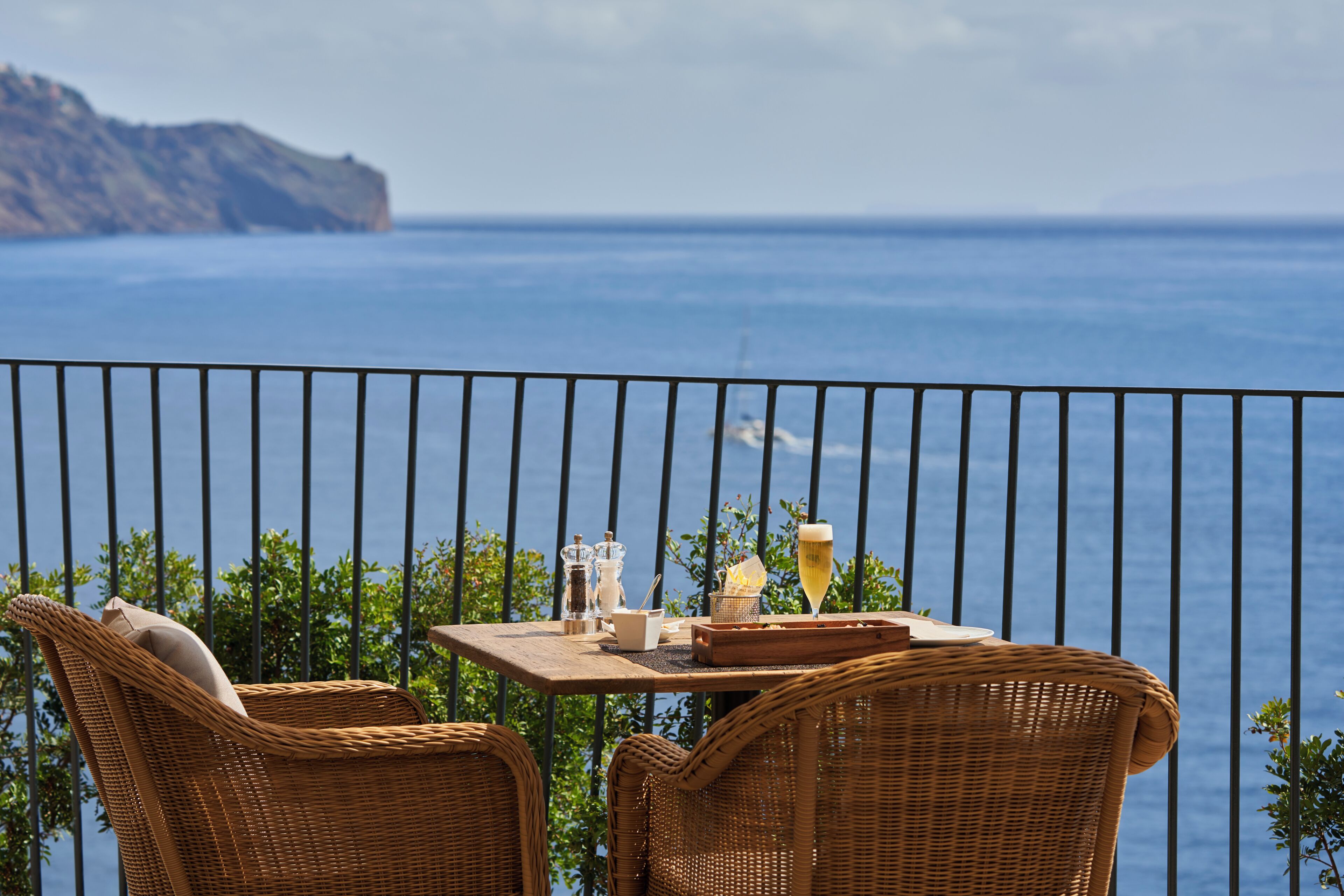 Varanda com cadeiras de vime, uma mesa com um copo de vinho e vista para o mar.