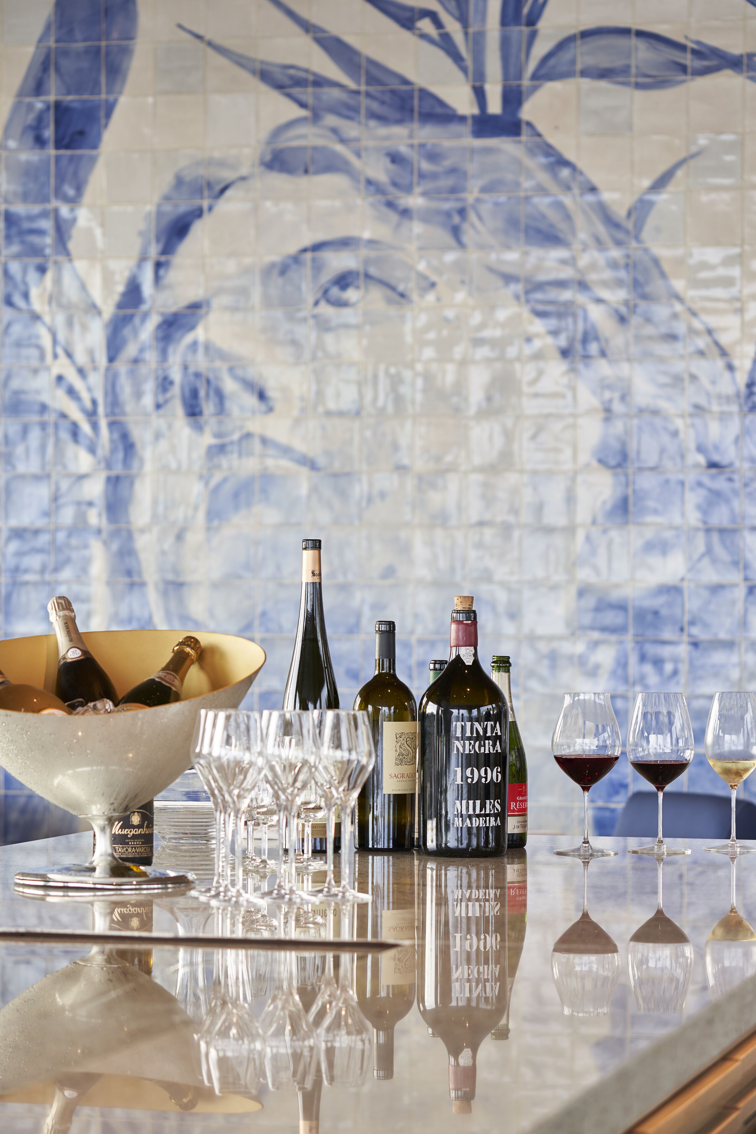 Una zona de bar con pared de azulejos, una copa de champán, botellas de vino y vasos sobre una superficie reflectante.