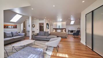 A spacious living area with a sofa, armchairs, and a coffee table. There is a bed in the background, a desk, and a wardrobe.