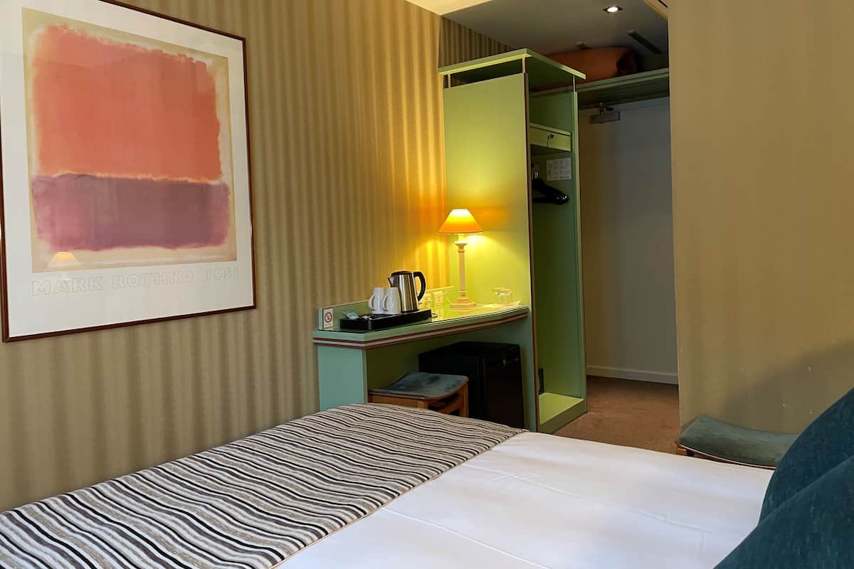 Una habitación de hotel con una cama, un escritorio con lámpara, una obra de arte enmarcada y un armario.