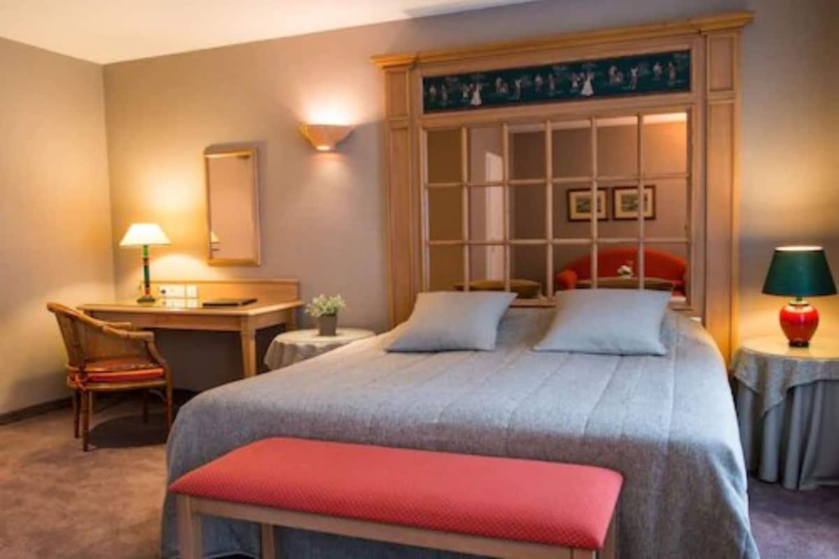 Habitación de hotel con cama, escritorio, silla, lámpara y una pequeña mesa con una planta.